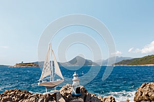 A wooden sailing ship with a lighthouse stand on a rocky shore against the sea. Against the backdrop of Fort Mamula in