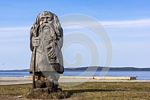 Wooden sculpture of old man in bast shoes with gusli and staff on lake shore