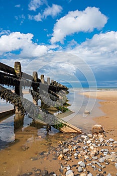 Wooden sea defence on the sandy beach, with blue sky and fluffy white clouds.