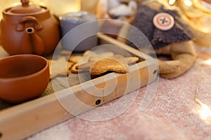 Wooden serving tray with brown ceramic kettle, steaming hot drink mug in a cozy home interior. Food, drinks, toy, gingerbread