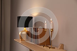 Wooden shelf with different books, burning candles and lamp on light wall