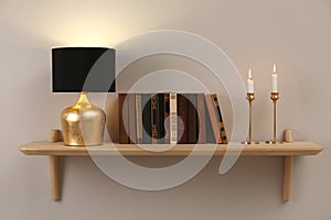 Wooden shelf with different books, burning candles and lamp on light wall