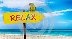 Wooden sign on beautiful beach and clear sea wit text relax. A green parrot sits on a pointer to a tropical paradise