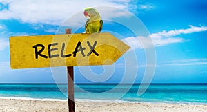 Wooden sign on beautiful beach and clear sea wit text relax. A green parrot sits on a pointer to a tropical paradise