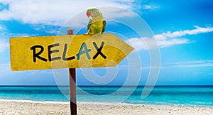 Wooden sign on beautiful beach and clear sea wit text relax. A green parrot sits on a pointer to a tropical paradise