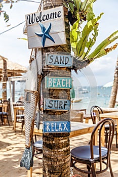 Wooden signs WELCOME, SAND, BEACH, SURF, RELAX and a wooden fish on a tree at a beach side cafe at Pantai Sanur, Bali, Indonesia.