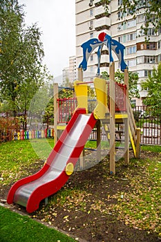 A wooden slide of bright red and yellow color against the background of green trees and residential buildings