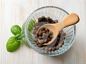 Wooden spoon with allspice jamaica pepper in a glass bowl and fresh basil leaves over white wooden table. Organic spice and