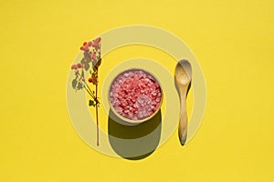 Wooden spoon and bowl filled with pink bath sea salt. Beauty treatment for spa and wellness on yellow background.