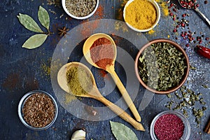 Wooden spoons with yellow and orange spices, different cups with dried herbs, garlic, bay leaf on a dark blue background