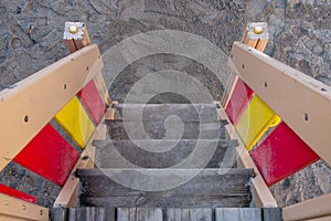 Wooden staircase on playground with plank steps covered with gray sand and red and yellow railings. Top view.