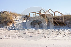 Wooden stairway on white sand beach with grass. Ocean coast landscape. Footpath on white sand and wooden outdoor stairs.