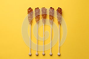 Wooden sticks with sugar crystals on yellow background, flat lay. Tasty rock candies