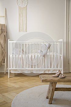 Wooden stool with shoes on round rug in bright baby`s bedroom interior with white cradle