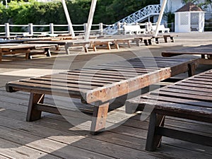 Wooden sun beds stand in the shade on a platform without a single vacationer next to the sea in the early summer morning.