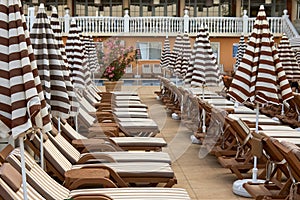 Wooden sunbeds with umbrellas for sunbathers stand in rows along the pool