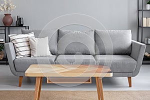 Wooden table and big grey couch with pillows in living room of trendy apartment, real photo