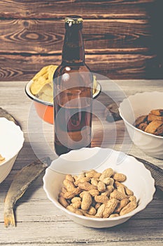 On a wooden table a bottle of beer, dried fish, chips, crackers and nuts