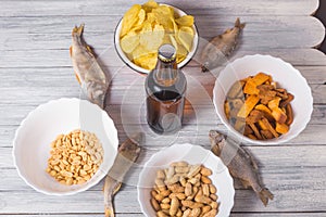 On a wooden table a bottle of beer, dried fish, chips, crackers and nuts