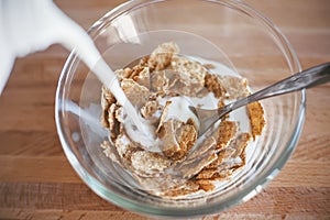 On a wooden table is a bowl with oatmeal cereals which is filled with milk. Breakfast in the early morning