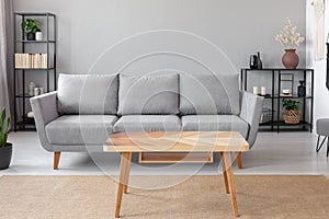 Wooden table on carpet in front of grey sofa in minimal living room interior with plant