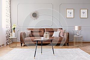 Wooden table on carpet in front of leather sofa in grey flat interior with posters and mirror