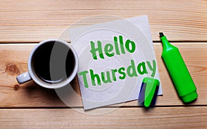 On a wooden table, a cup of coffee, a green marker and a napkin with the text HELLO THURSDAY