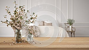 Wooden table, desk or shelf close up with branches of cherry blossoms in glass vase over blurred view of classic bedroom with soft