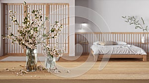 Wooden table, desk or shelf close up with branches of cherry blossoms in glass vase over blurred view of japandi bedroom,