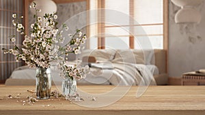 Wooden table, desk or shelf close up with branches of cherry blossoms in glass vase over wooden bedroom with double bed, japandi
