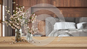 Wooden table, desk or shelf close up with branches of cherry blossoms in glass vase over wooden minimal bedroom with double bed,