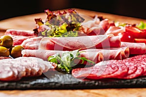 Wooden table filled with delicious cold meat selection. A plate with a variety of sausages with green vegetables green lettuce,