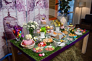 A wooden table with a grassy tablecloth and Easter treats: eggs, cakes, chocolate eggs, meringues