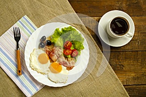 On a wooden table on a linen napkin, a plate with fried eggs with bacon and a cup of coffee.