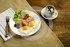 On a wooden table on a linen napkin, a plate with fried eggs with bacon and a cup of coffee.