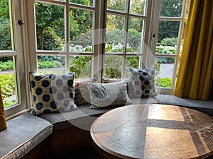 Wooden table near cozy bay window seat with cushions in room. Interior design