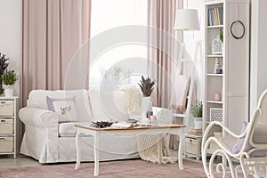 Wooden table next to white couch in pink living room interior with drapes and plants. Real photo