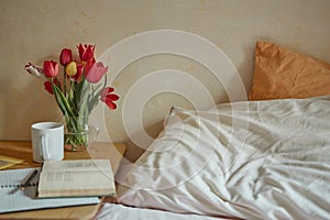 Wooden table with paper notebook, book, cup of tea and spring flowers next to bed