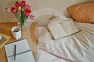 Wooden table with paper notebook, book, cup of tea and spring flowers next to bed
