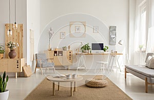 Wooden table and pouf on carpet in white living room interior with poster and workspace. Real photo