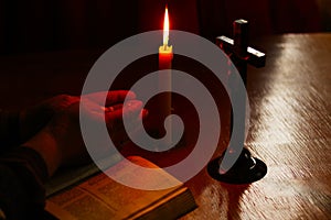 Wooden table in retro style. On it is a candle and a cross. Hands of an old woman over a book. Palms folded