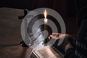 Wooden table in retro style. On it is a candle and a cross. Hands of an old woman over a book. Palms folded