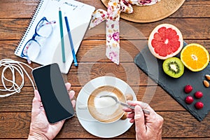 Wooden table set for an healthy breakfast. Hot cappuccino and citrus fruit. Human hand holds a cellphone