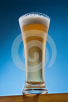 On a wooden table, a tall glass is full of beer with foam, on a blue background, close-up