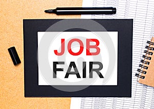 On a wooden table, there is a black open marker, a brown notepad and a sheet of paper in a black frame with the text JOB FAIR.