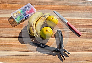 On a wooden table there are a multitool, a knife, a box of fishing tackle and fruits