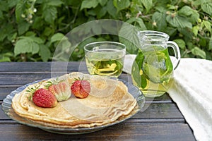 On a wooden table there is a plate with pancakes on which there are several strawberries, next to a transparent pitcher