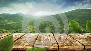 Wooden table top with green mountain background. Blue sky with sun light and green small leaves.