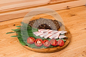 On the wooden table top is a round Board with sliced black bread with seeds, smoked brisket, tomatoes and herbs photo