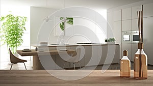 Wooden table top or shelf with aromatic sticks bottles over blurred modern white kitchen with parquet floor and island with stools